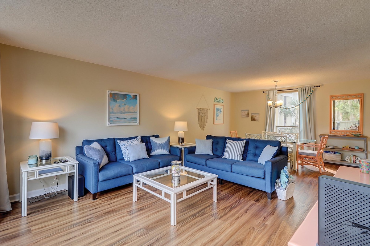 A spacious living area is presented with comfortable blue sofas and a coordinating coffee table. Natural light enhances the neutral walls, complemented by light wood flooring. An adjacent dining space features a round table with four chairs, creating an inviting atmosphere for gatherings.