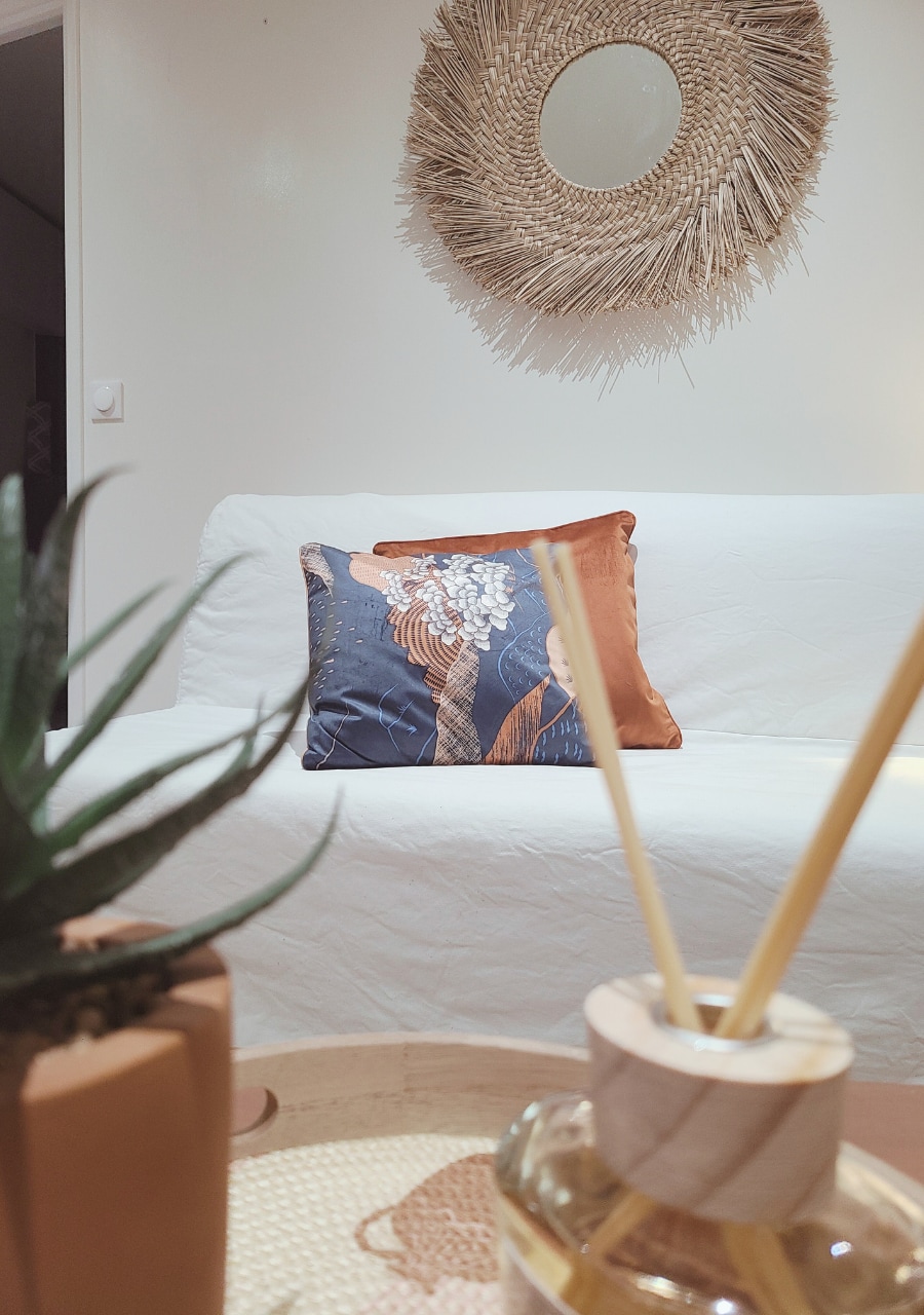A neutral-toned sofa is adorned with a decorative pillow featuring floral designs. A round mirror with a natural woven frame hangs above, reflecting light. A plant and diffuser with wooden accents sit on a tray in the foreground, enhancing the cozy and stylish ambiance.