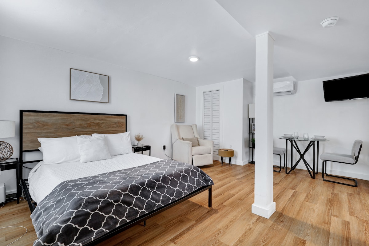 The studio features a spacious sleeping area with a queen-sized bed, complemented by a recliner and a bedside table. Natural light illuminates the space, showcasing a comfortable and minimalistic design. A smart TV is mounted on the wall, adding convenience for entertainment.