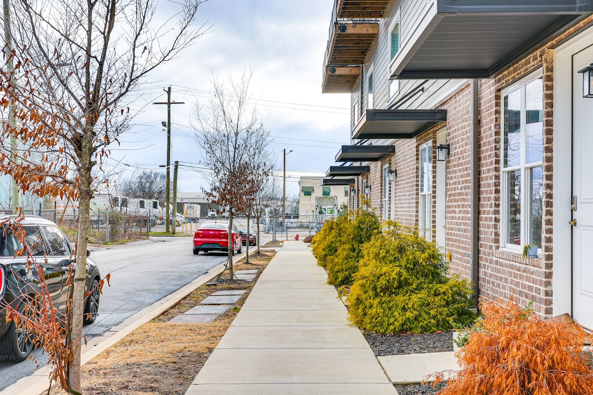 A well-maintained sidewalk lines the exterior of a modern building with brick and siding. Neatly trimmed shrubs and small trees enhance the landscape. Parked vehicles are visible along the street, and the sky is overcast.
