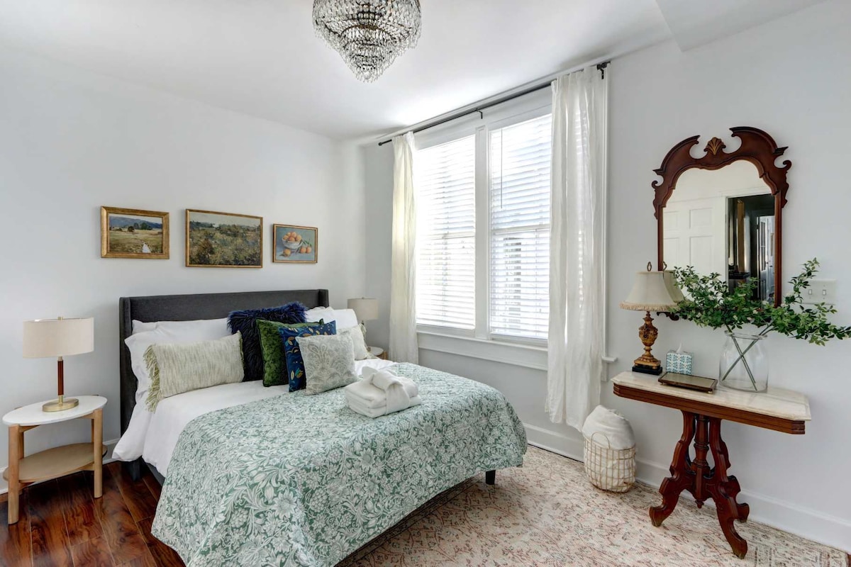 A serene bedroom features a queen bed dressed in soft linens with a decorative green and white quilt. Two bedside lamps illuminate the space, alongside a vintage mirror and a small table adorned with a plant. Natural light filters through the sheer curtains of the large windows.