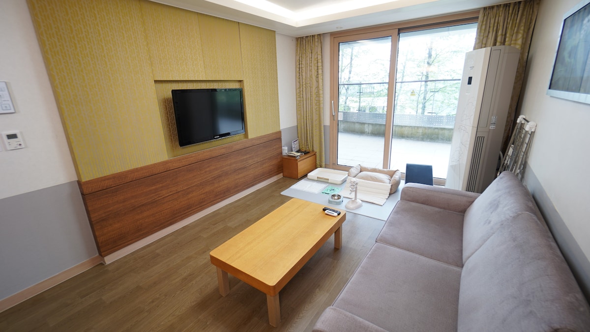 A spacious living area features a comfortable sofa in a neutral tone. A wooden coffee table sits in front, with a flat-screen TV mounted on the wall. Light streams in from large glass doors, leading to a balcony with greenery visible outside.