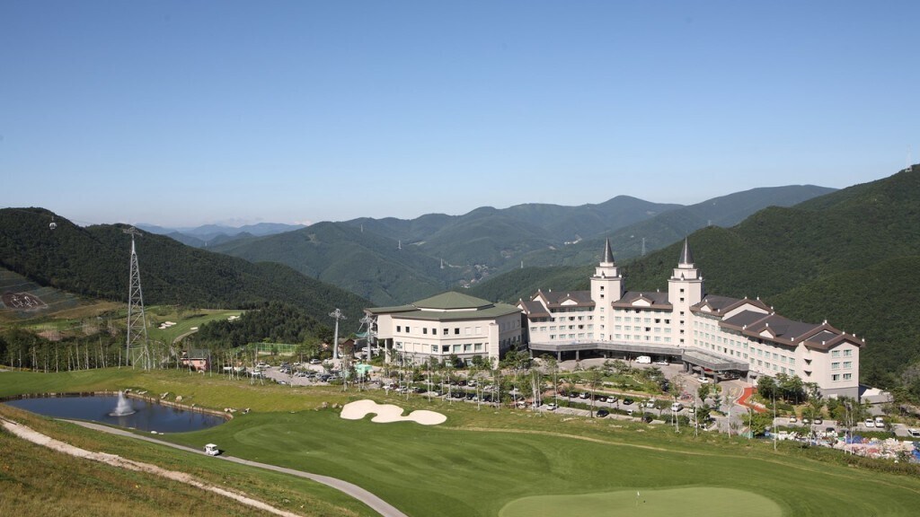 The building features a distinctive architectural style, with two prominent turrets. It is surrounded by lush green hills and a golf course, providing a tranquil outdoor setting. A peaceful pond stretches in the foreground, reflecting the landscape under a clear blue sky.