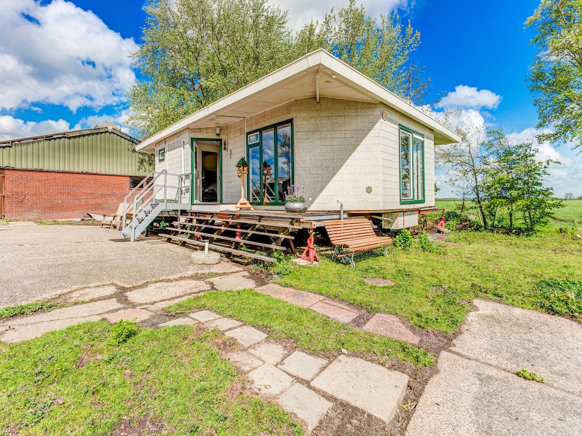 A modern single-story home is set on stilts, surrounded by greenery and open space. Large glass doors provide natural light and a view of the exterior. A simple pathway of stones leads from the driveway to the entrance, enhancing accessibility to the home.