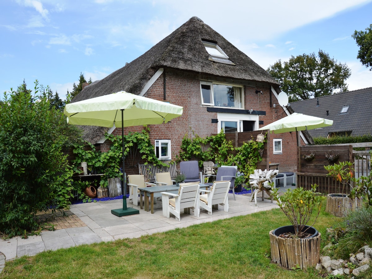 A charming garden area is outlined by lush greenery, featuring multiple patio tables and chairs under large umbrellas. The rustic brick farmhouse stands in the background, complemented by the relaxing outdoor setting and decorative planters.