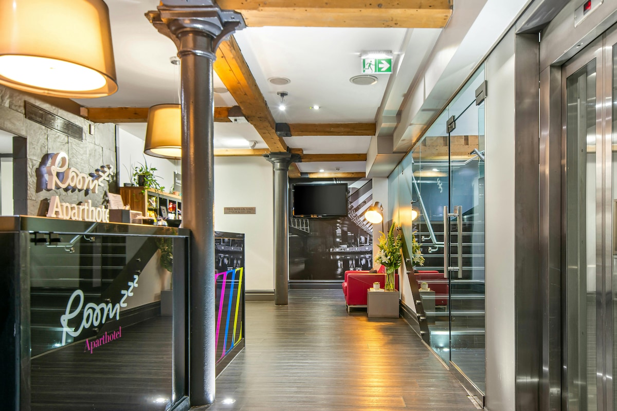 The lobby area features a blend of modern and historic design, with exposed wooden beams and stylish columns. A reception desk is situated to the left, alongside comfortable seating areas, where a red chair adds a pop of color. Soft lighting creates a welcoming atmosphere.