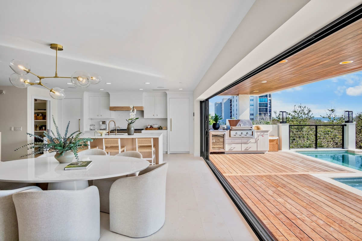 An open-concept dining area features a modern round table with soft, upholstered chairs. Bright natural light floods the space through expansive sliding glass doors that lead to a wooden deck, showcasing an outdoor kitchen and a glimpse of the infinity pool beyond.