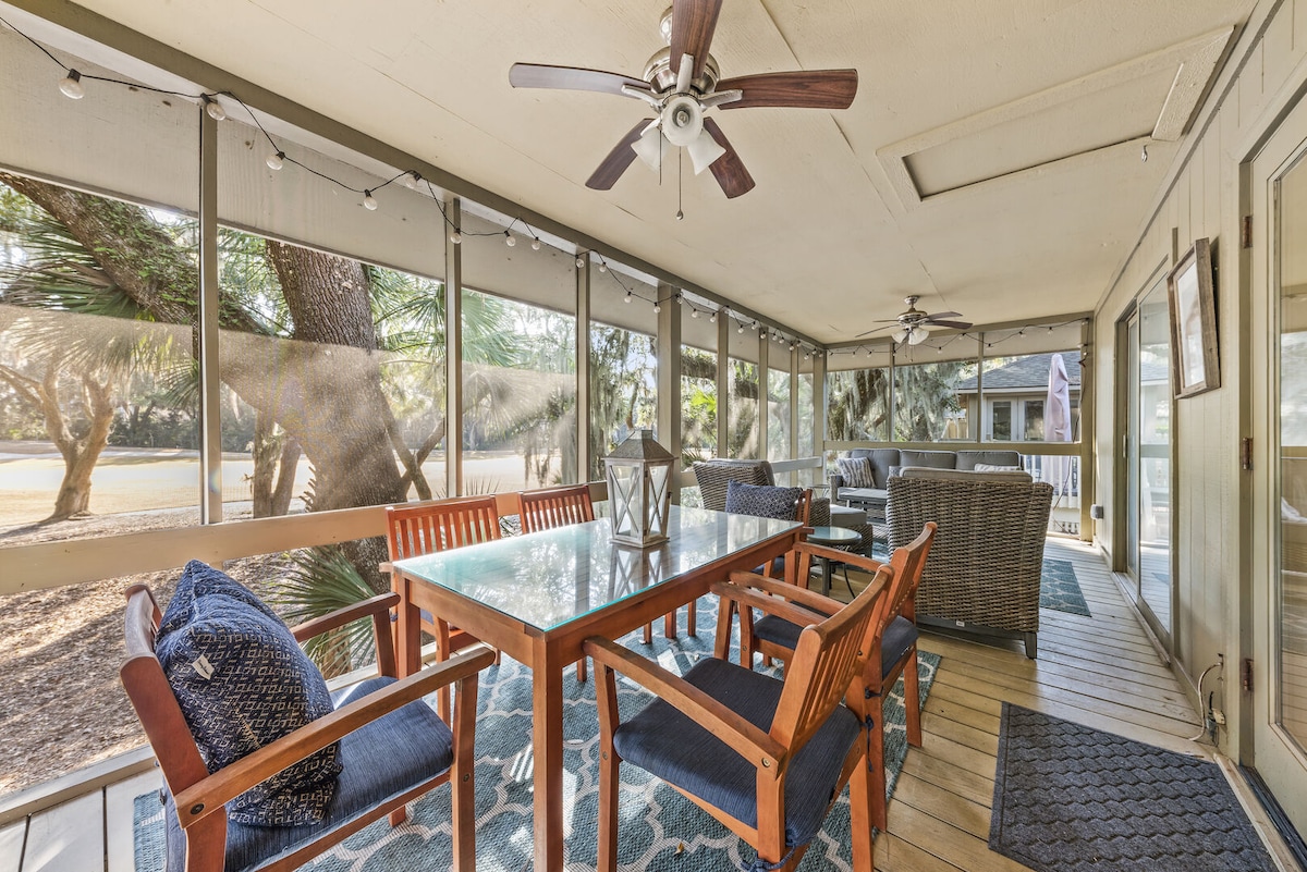A spacious screened porch features a large wooden dining table surrounded by several chairs, ideal for outdoor meals. Comfortable seating is arranged nearby, including a cozy sectional. Ceiling fans provide airflow, while views of the surrounding greenery and fairway create a peaceful atmosphere.