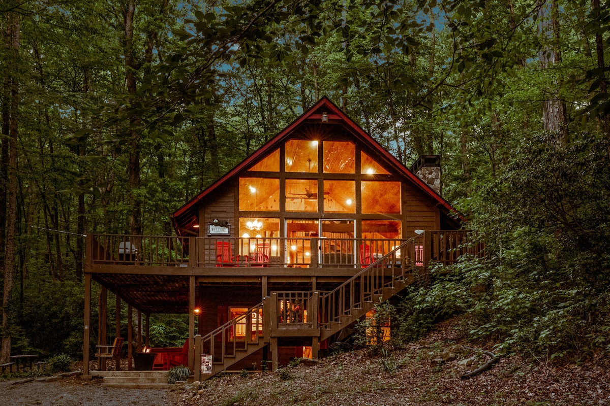 A cabin nestled among trees features a wraparound deck and large windows that illuminate the interior. The two-story structure blends rustic charm with modern design, inviting guests to enjoy the surrounding nature. Warm lighting emits a cozy ambiance from within.