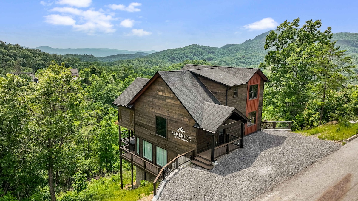 A modern cabin is nestled among verdant mountains, featuring a combination of wood siding and large windows. The property is surrounded by lush greenery, showcasing a spacious outdoor area and scenic views of the surrounding hills under a clear blue sky.