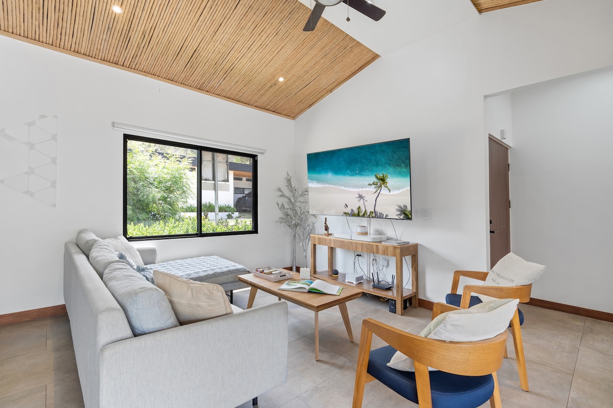 A bright living area features a comfortable sectional sofa next to a natural wood coffee table. A large television is mounted on the wall, while a ceiling fan and bamboo ceiling add a touch of warmth to the space. Expansive windows open to greenery outside.