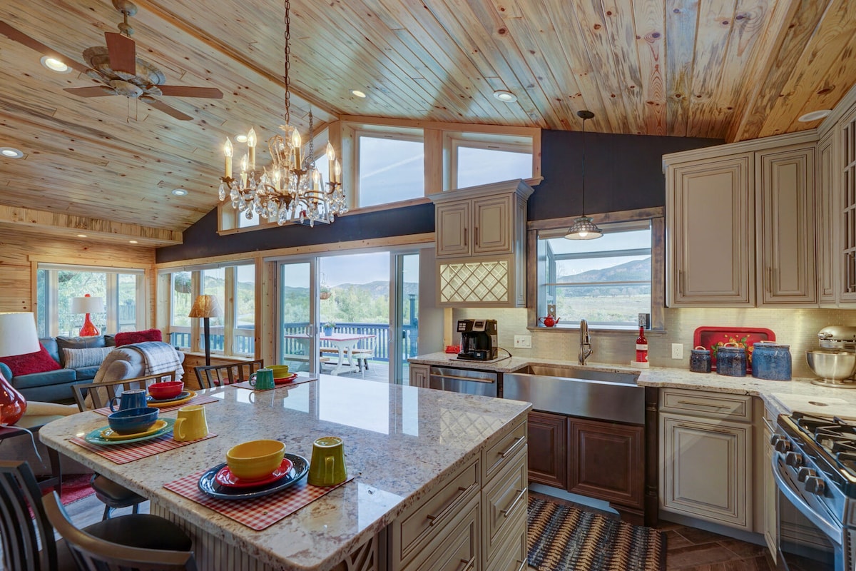 A spacious kitchen is presented, featuring a large central island with colorful dishware arranged atop. Sunlight pours through multiple windows, illuminating the wooden ceiling and cabinetry. Cozy seating is visible in the background, enhancing a warm gathering space.
