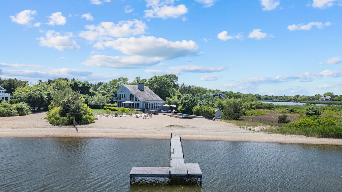 A spacious beachfront home is set against a bright blue sky, with lush greenery surrounding the property. A private dock extends into the calm water, while lounge chairs are positioned on the sandy shore, inviting outdoor relaxation and enjoyment of the scenic views.