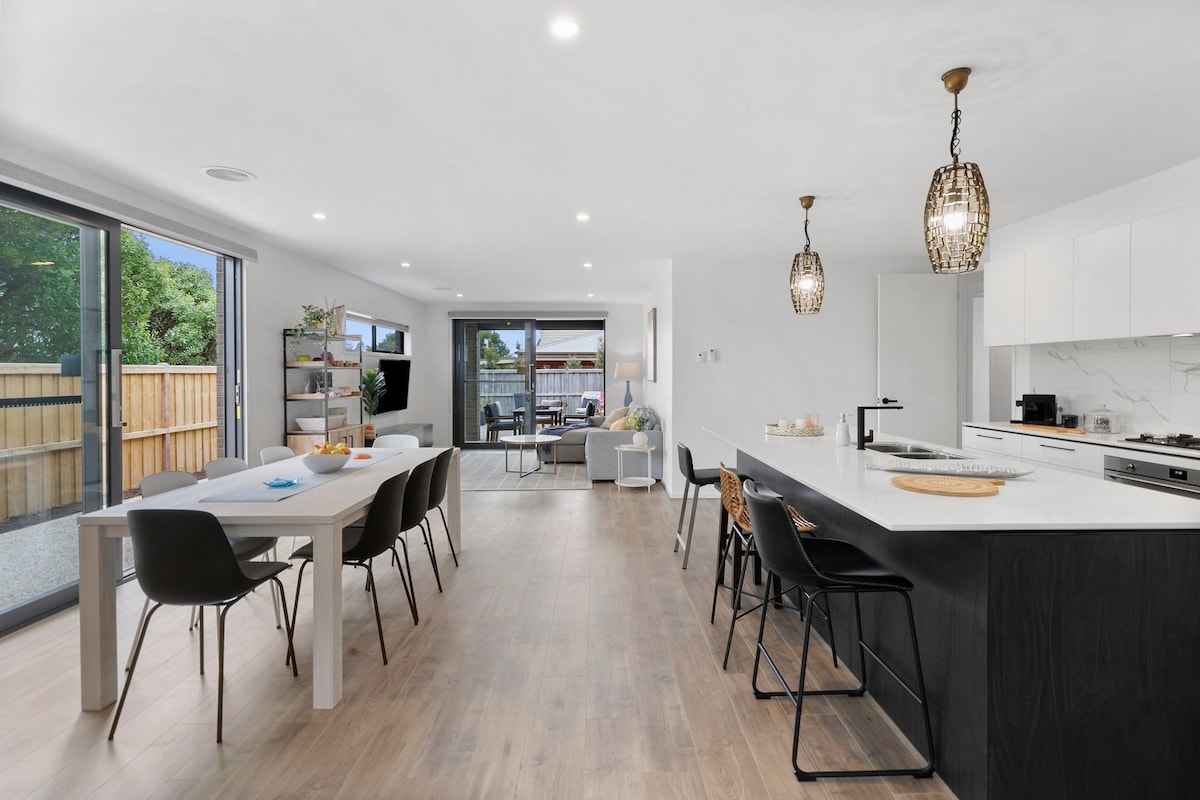 An open-plan living area features a modern kitchen with a large stone island and stylish pendant lights. A dining table with eight chairs is positioned nearby. Large windows allow natural light to fill the space, with views of the fenced backyard visible through sliding glass doors.