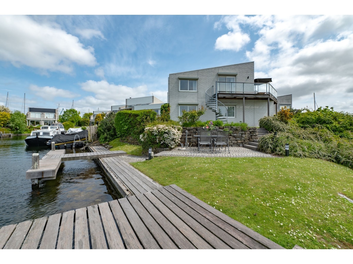 The water villa is situated by the edge of the lake, featuring a private jetty and well-maintained garden. A spiral staircase leads to an upper balcony, adding dimension to the outdoor space. Comfortable seating is positioned along the water's edge, surrounded by lush greenery.