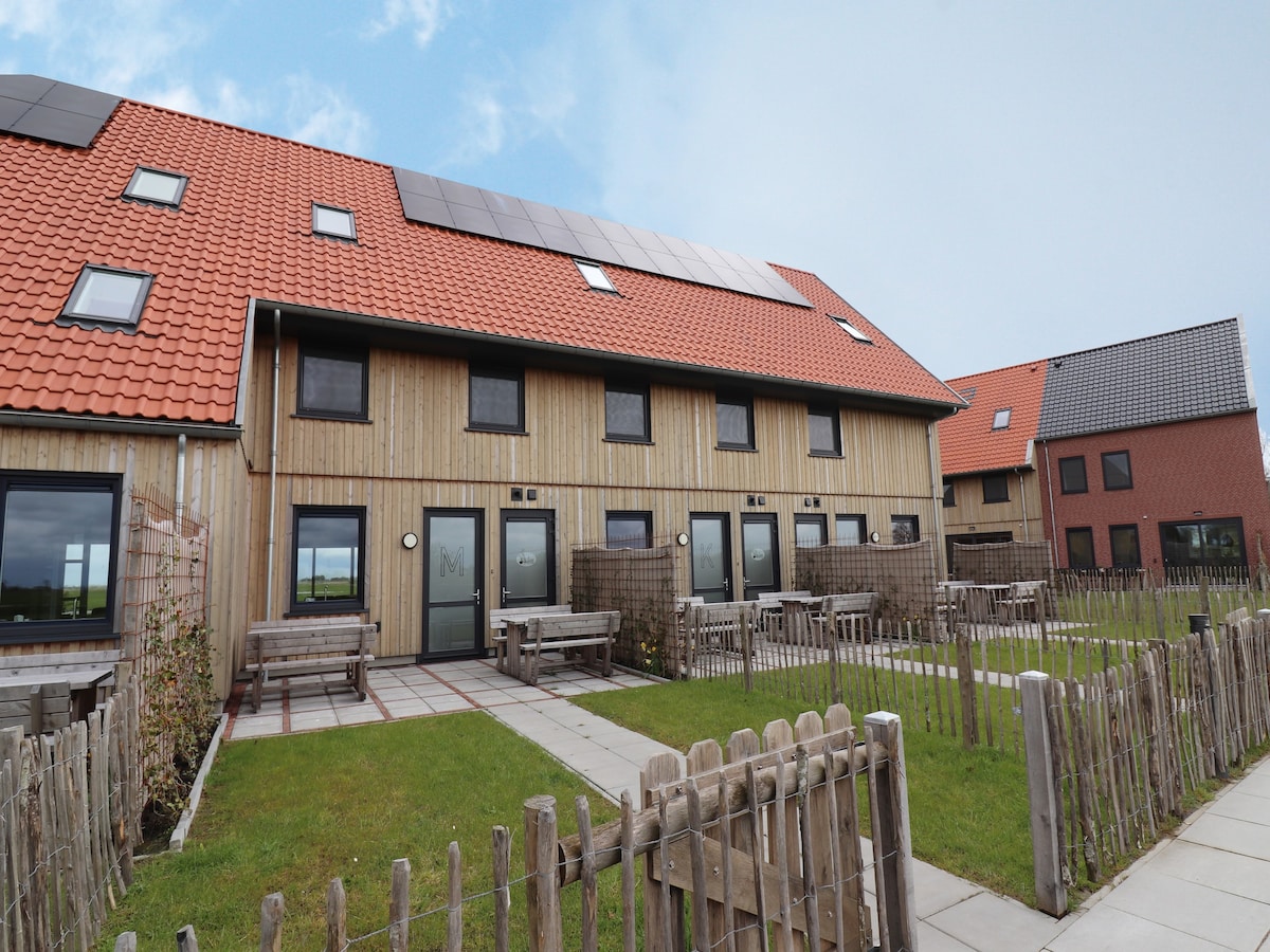 The exterior of the villa features a wooden facade with a red-tiled roof, large windows, and solar panels. A pathway leads through the grassy area, accompanied by wooden benches and a fenced garden space, creating a welcoming environment for relaxation.