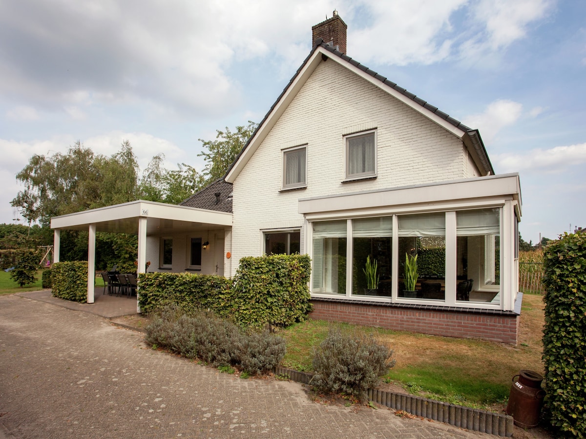 The exterior of a two-story cottage features a white façade with large glass windows and a covered porch. Surrounding greenery includes well-maintained hedges and a grassy lawn. A gravel pathway leads up to the entrance, complemented by a clear blue sky and scattered clouds.