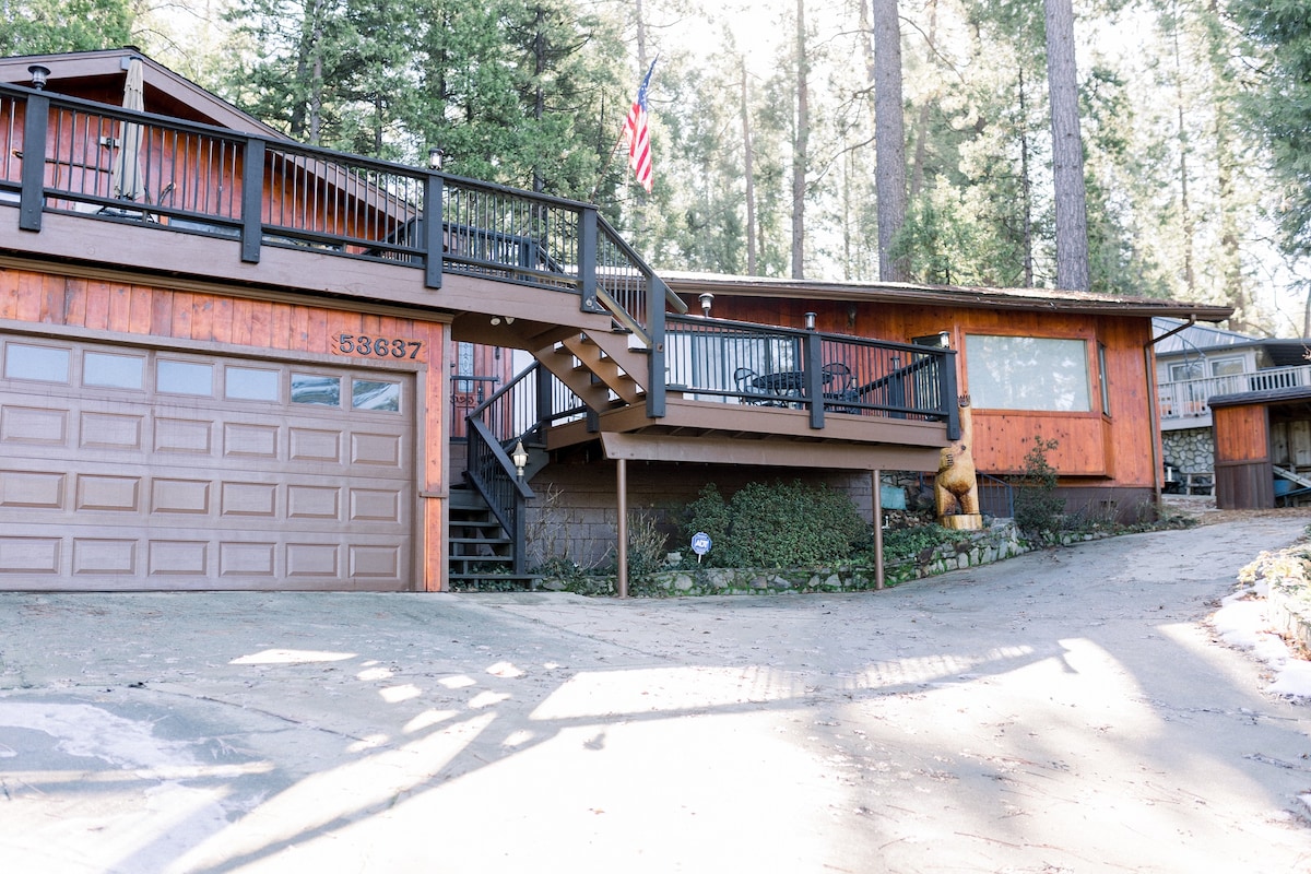 An exterior view of the home showcases a wooden structure with large windows and a multi-level deck. The American flag is displayed prominently. A paved driveway leads to a garage, and surrounding pine trees add to the natural setting.