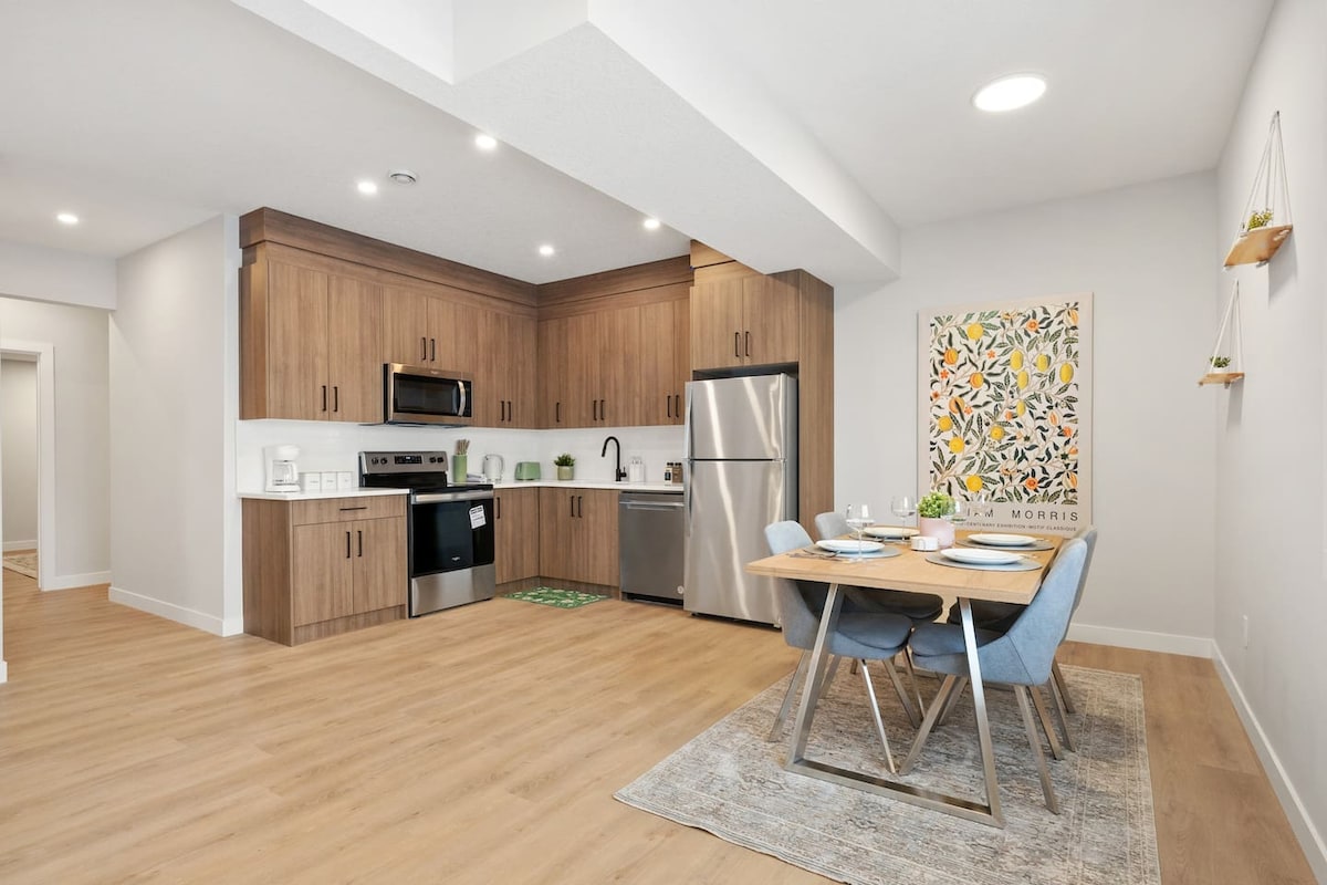 A modern kitchen and dining area feature light wood cabinetry and stainless steel appliances. A dining table with four blue chairs is set for a meal, complemented by a colorful wall art piece. The light wood flooring and warm lighting create a welcoming ambiance.