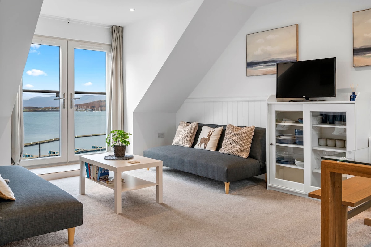 A bright living area features two comfortable gray sofas, a light coffee table, and a glass-fronted cabinet. Floor-to-ceiling windows provide views of the sea and surrounding landscape. Soft neutral tones and natural light create a peaceful setting for relaxation.