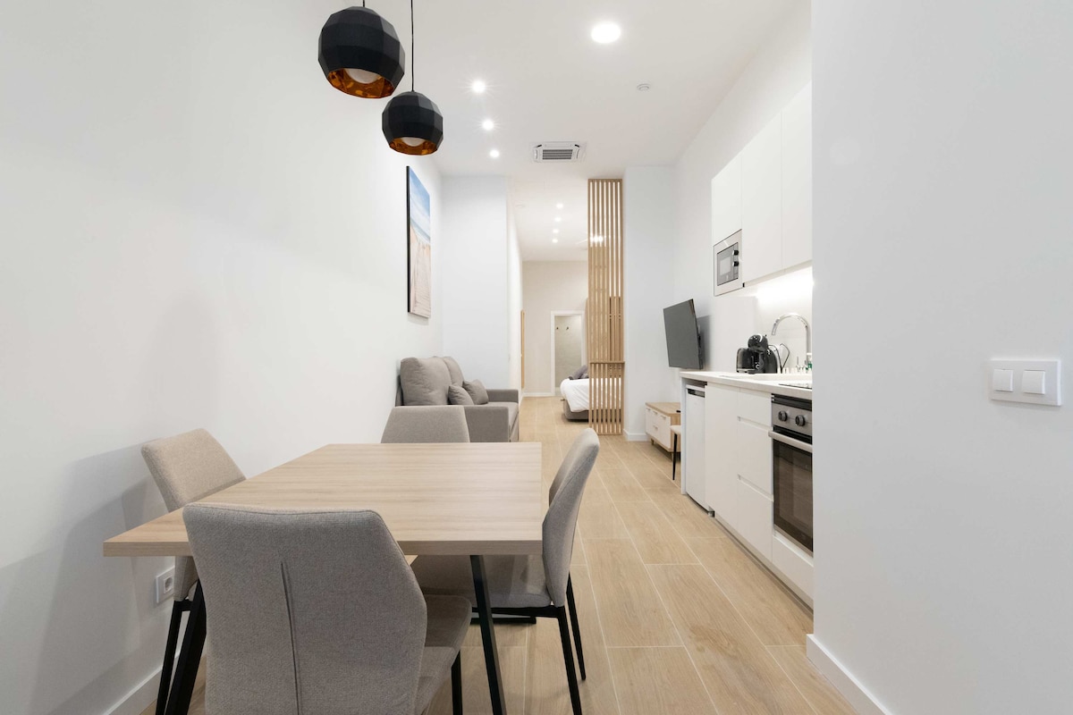 An open-plan living area features a wooden dining table surrounded by four upholstered chairs. In the background, a comfortable sitting area with soft furnishings is visible. The kitchen area is equipped with modern appliances and cabinets, enhancing the contemporary design.