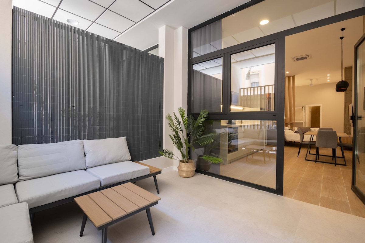 A comfortable seating area features a light-colored sofa and a wooden coffee table. A potted plant adds a touch of greenery, while large glass doors provide a view into the interior space, which is illuminated by soft lighting.