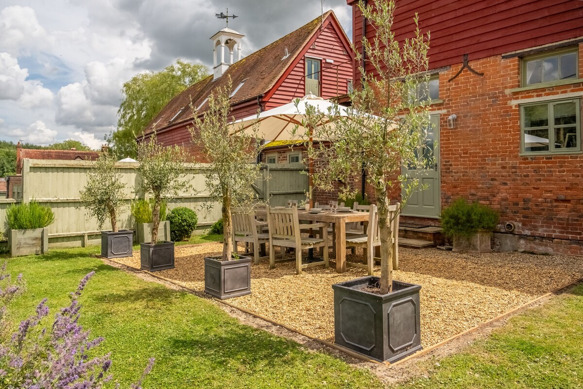 An outdoor space featuring a gravelled area with a wooden dining table and chairs, shaded by a large umbrella. Potted olive trees are placed around the space, adding greenery. The surrounding garden is well-maintained, providing a serene setting for outdoor dining.