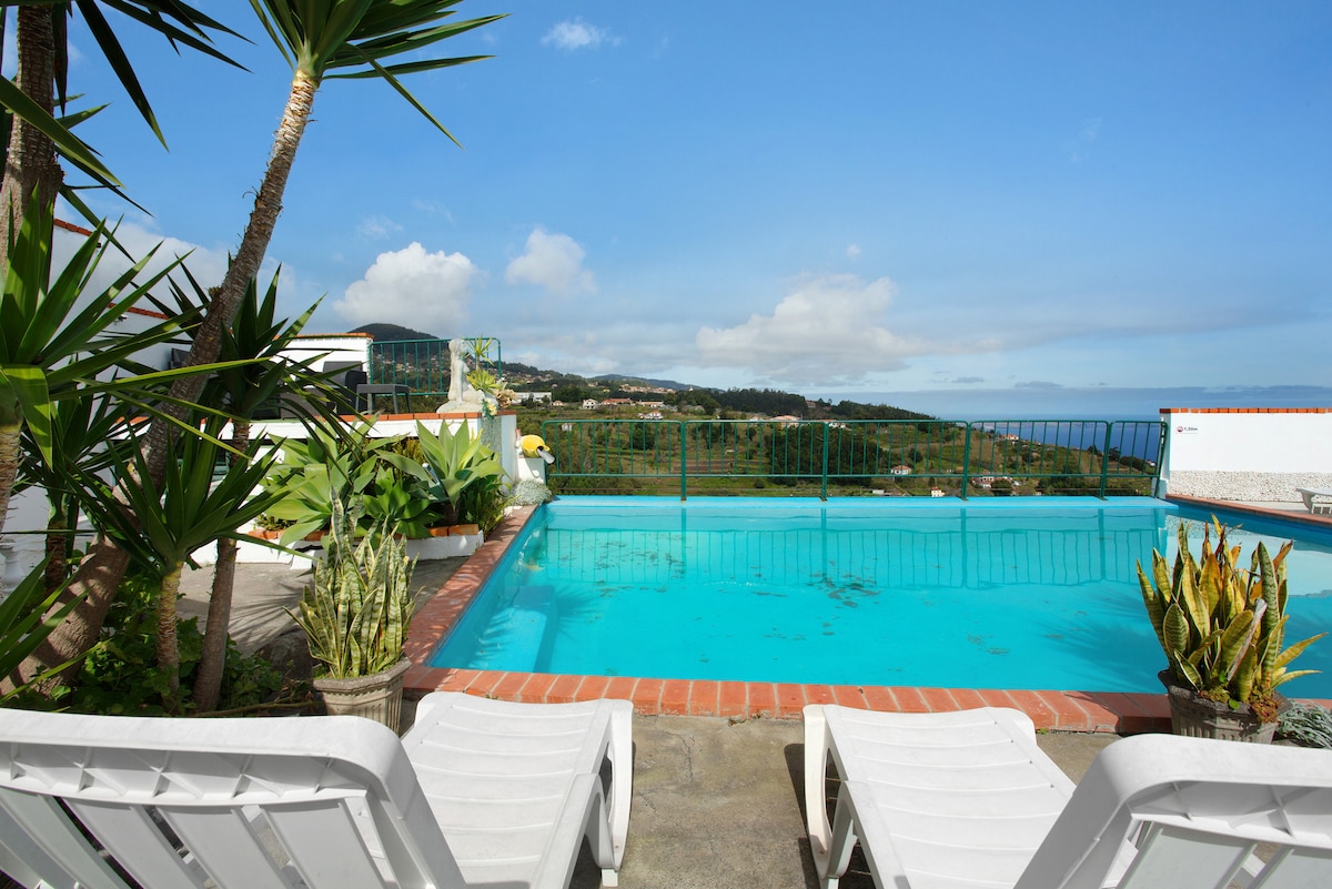 A sunny outdoor pool area is presented, featuring a turquoise pool surrounded by potted plants and lounge chairs. The view extends over hills and valleys toward the Atlantic, under a clear blue sky with a few clouds.