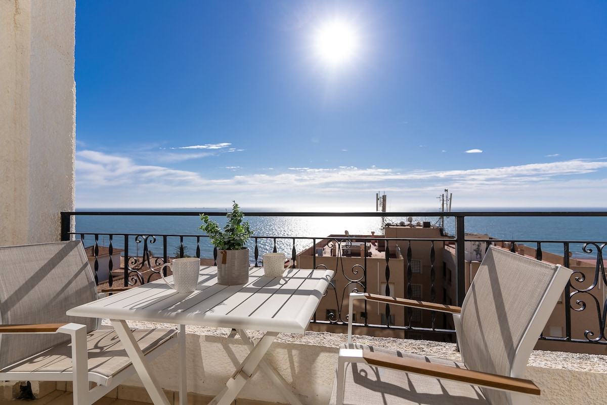 A balcony with two light-colored chairs and a white table is visible, overlooking the sea. A small plant is placed on the table. The sun shines brightly in a clear blue sky, creating a serene atmosphere.