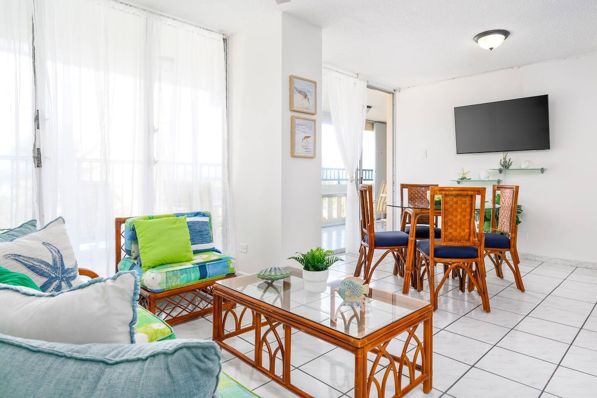 A bright living and dining area is presented, featuring light-colored tiled flooring. Woven rattan furniture is arranged with colorful cushions. A dining table is set for four, while a television is mounted on the wall nearby. Sheer white curtains frame the large windows, allowing natural light to fill the space.