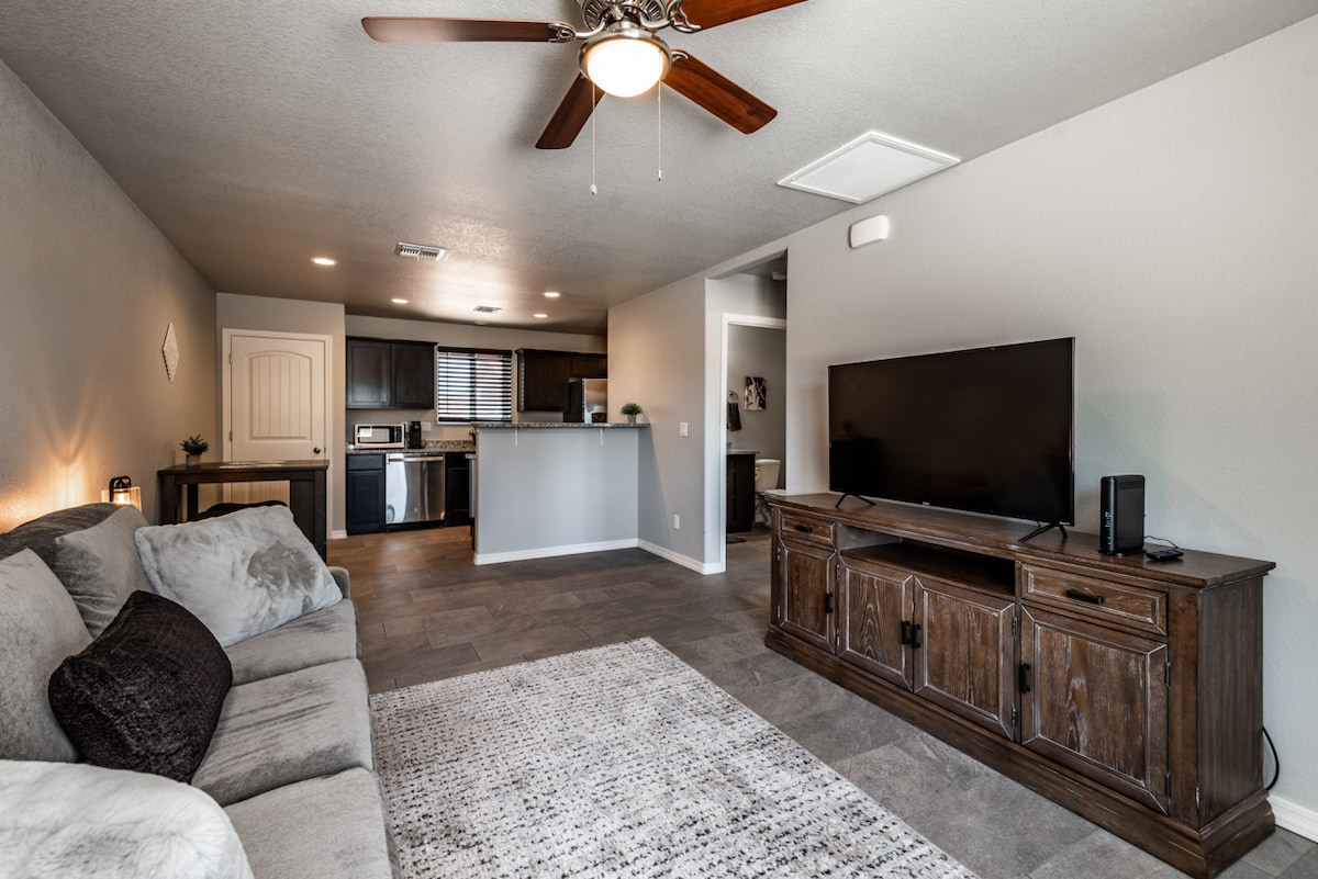 An open living area features a neutral-colored sectional couch with plush cushions, a wooden entertainment center housing a large television, and a ceiling fan. The contemporary kitchen, visible in the background, includes sleek cabinetry and modern appliances, complemented by natural light streaming through the windows.