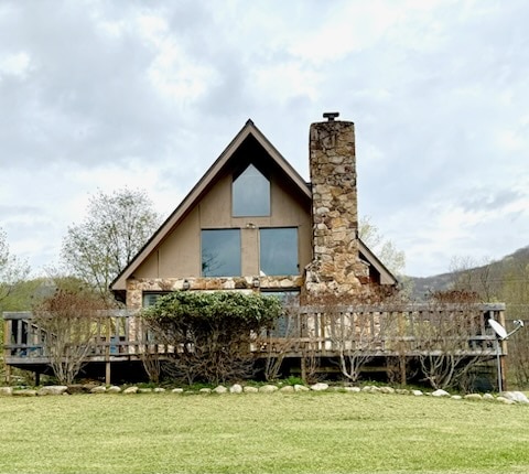 The exterior of a quaint cottage is highlighted, featuring a stone chimney and large glass windows. A wooden deck is visible, surrounded by low shrubs and manicured grass. The natural landscape offers a serene backdrop, enhancing the connection to the outdoors.