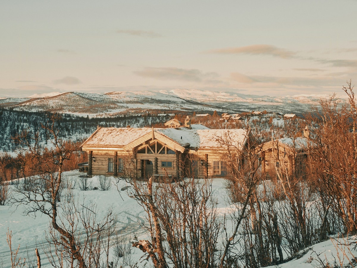 A scenic log cabin nestled in a winter landscape, surrounded by snow-covered hills and trees. The warm light of sunset casts a soft glow on the cabin's wooden exterior, highlighting its architectural features against the backdrop of distant mountains.