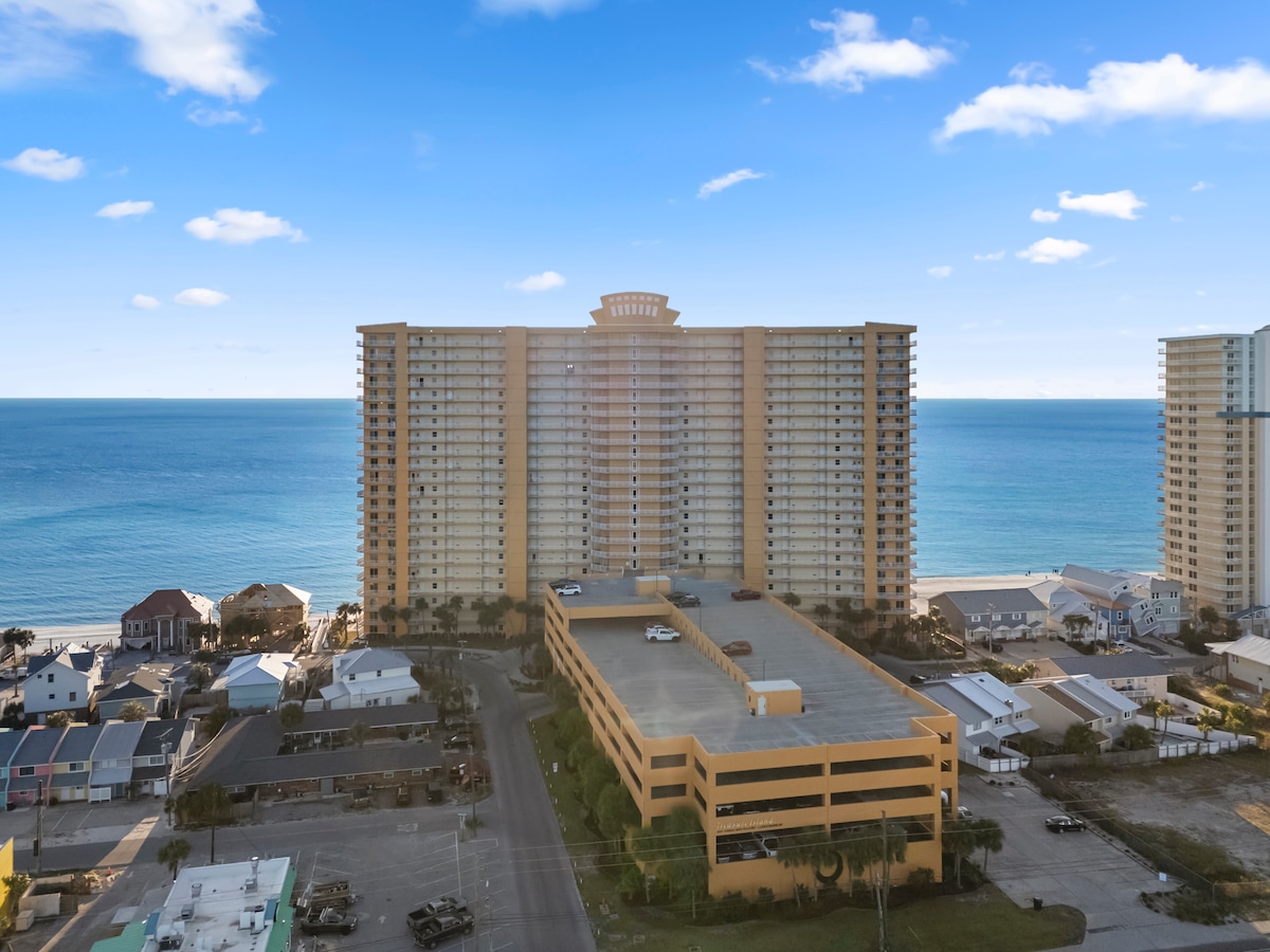 An aerial view of the Treasure Island Resort displays a towering structure overlooking the sparkling emerald waters of Panama City Beach. The nearby parking facility and beachside homes are visible, illustrating the proximity to lively beachfront attractions.