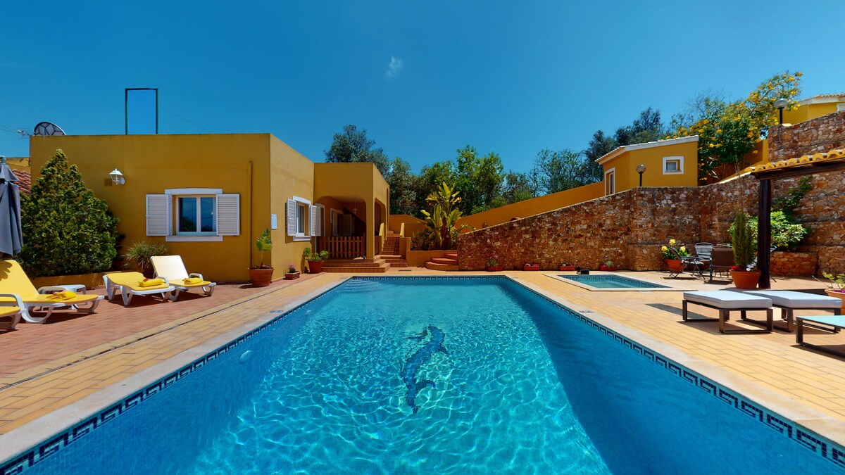 A spacious outdoor area features a large swimming pool surrounded by lounge chairs. A jacuzzi is visible to one side, with stone walls and lush greenery bordering the space. The bright yellow walls of the house complement the clear blue sky.
