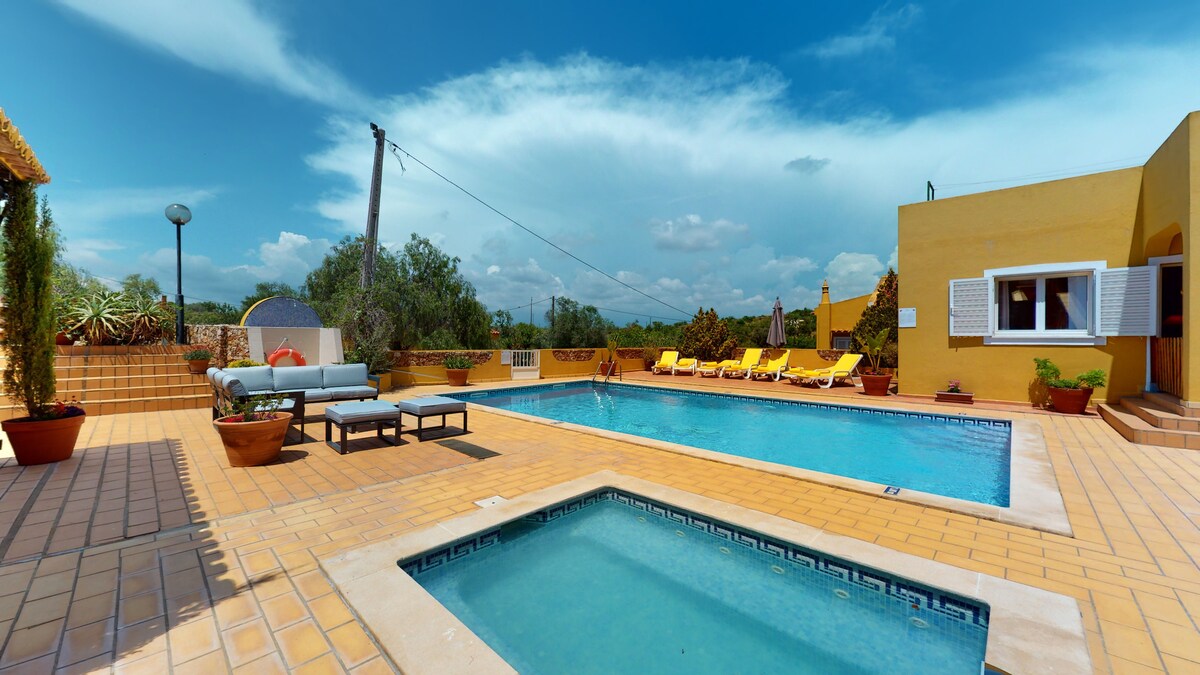 A spacious outdoor area features a shimmering pool surrounded by lounge chairs, with a separate jacuzzi nearby. Potted plants add greenery to the space. The clear blue sky and distant trees create a serene backdrop, ideal for relaxation.