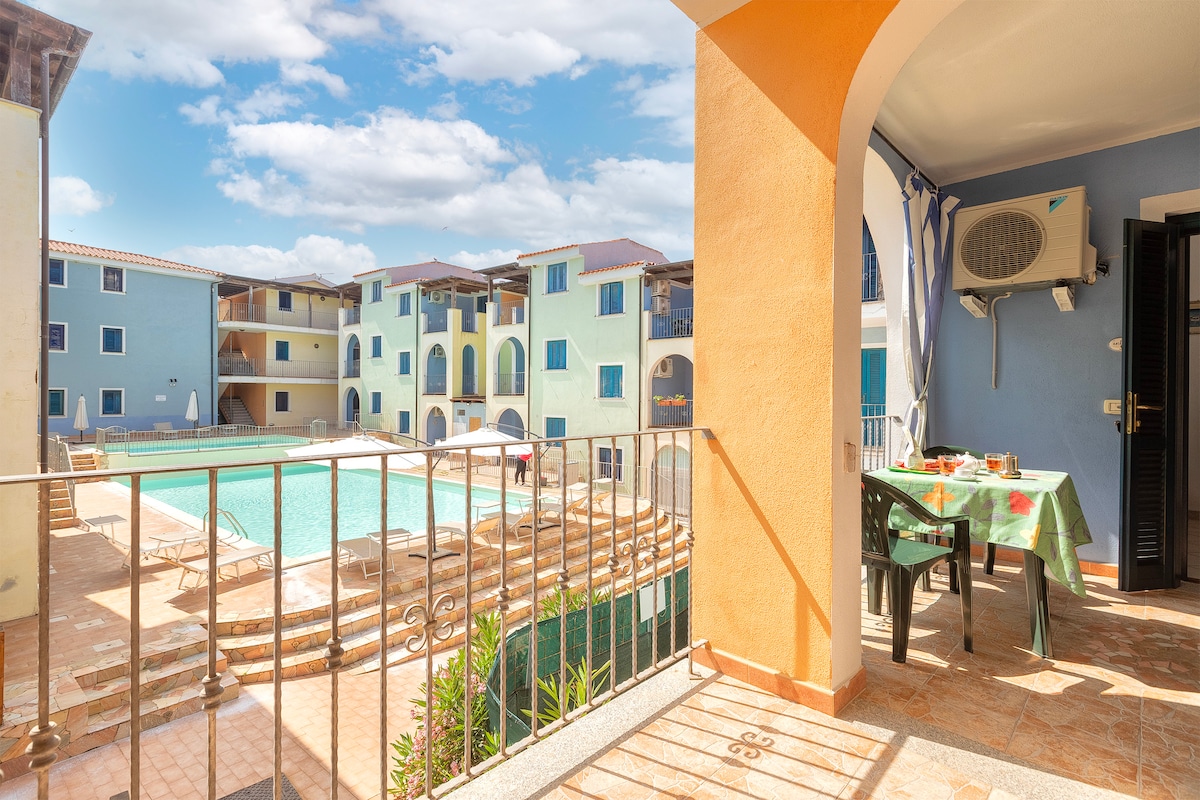 A private terrace is visible, featuring a table set for dining. A view of the shared outdoor area showcases a fenced pool surrounded by lounge chairs and colorful buildings, all under a bright sky with scattered clouds.