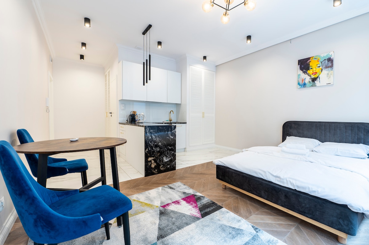 The spacious living area includes a comfortable double bed and a kitchenette with modern fixtures. A small dining table with two blue chairs is positioned nearby on a patterned rug. Soft lighting from stylish fixtures enhances the design of the room.