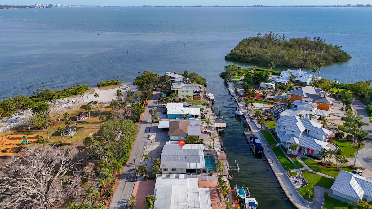 An aerial view captures the surrounding neighborhood by Sarasota Bay, featuring tree-lined streets and waterways. A mix of residential homes is visible, along with green parks and recreational areas, demonstrating the serene coastal environment a short distance from the water.
