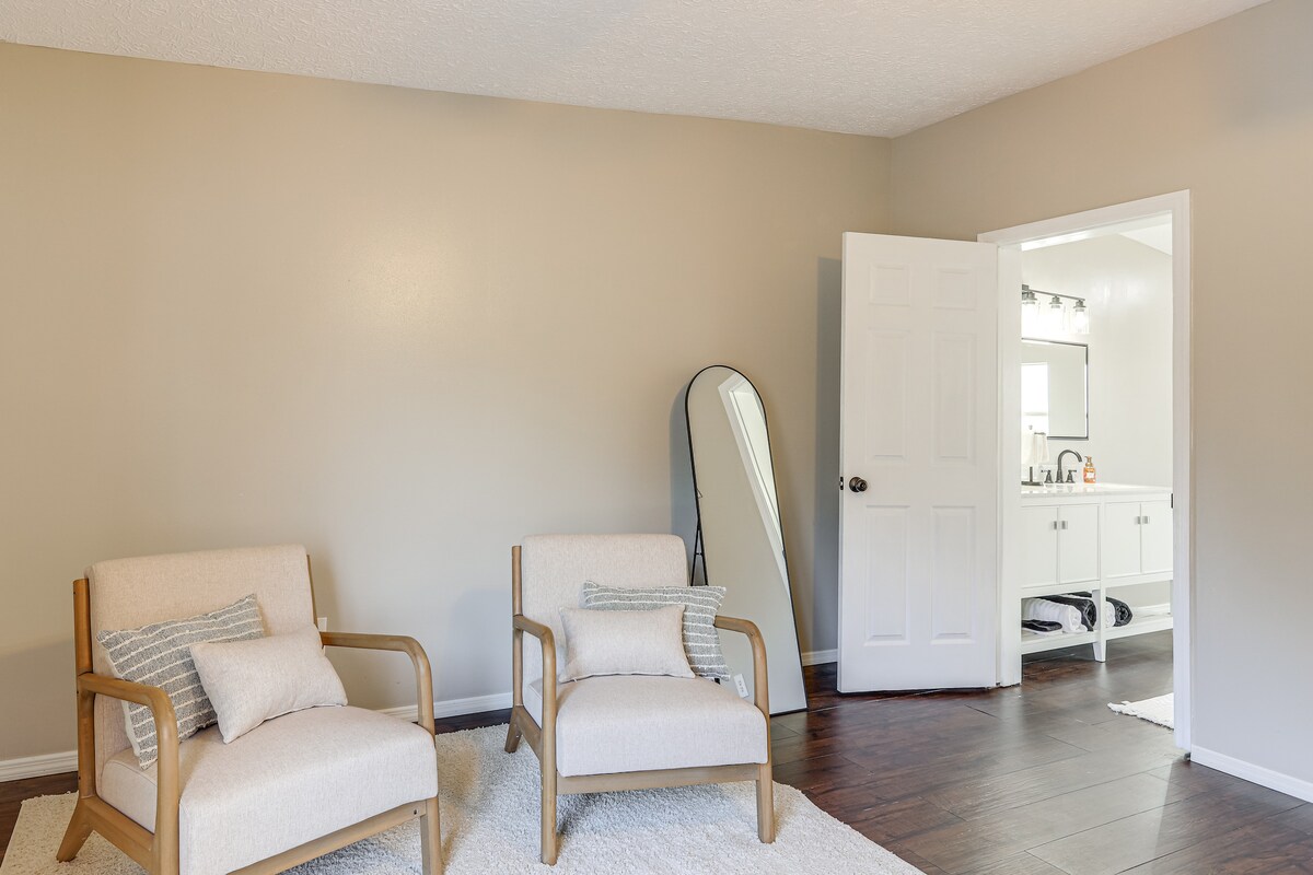 A spacious and neutral-toned sitting area features two wooden chairs with light upholstery and small accent pillows. A full-length mirror leans against the wall near an open doorway, which leads to a well-lit bathroom. Soft, natural light brightens the room.