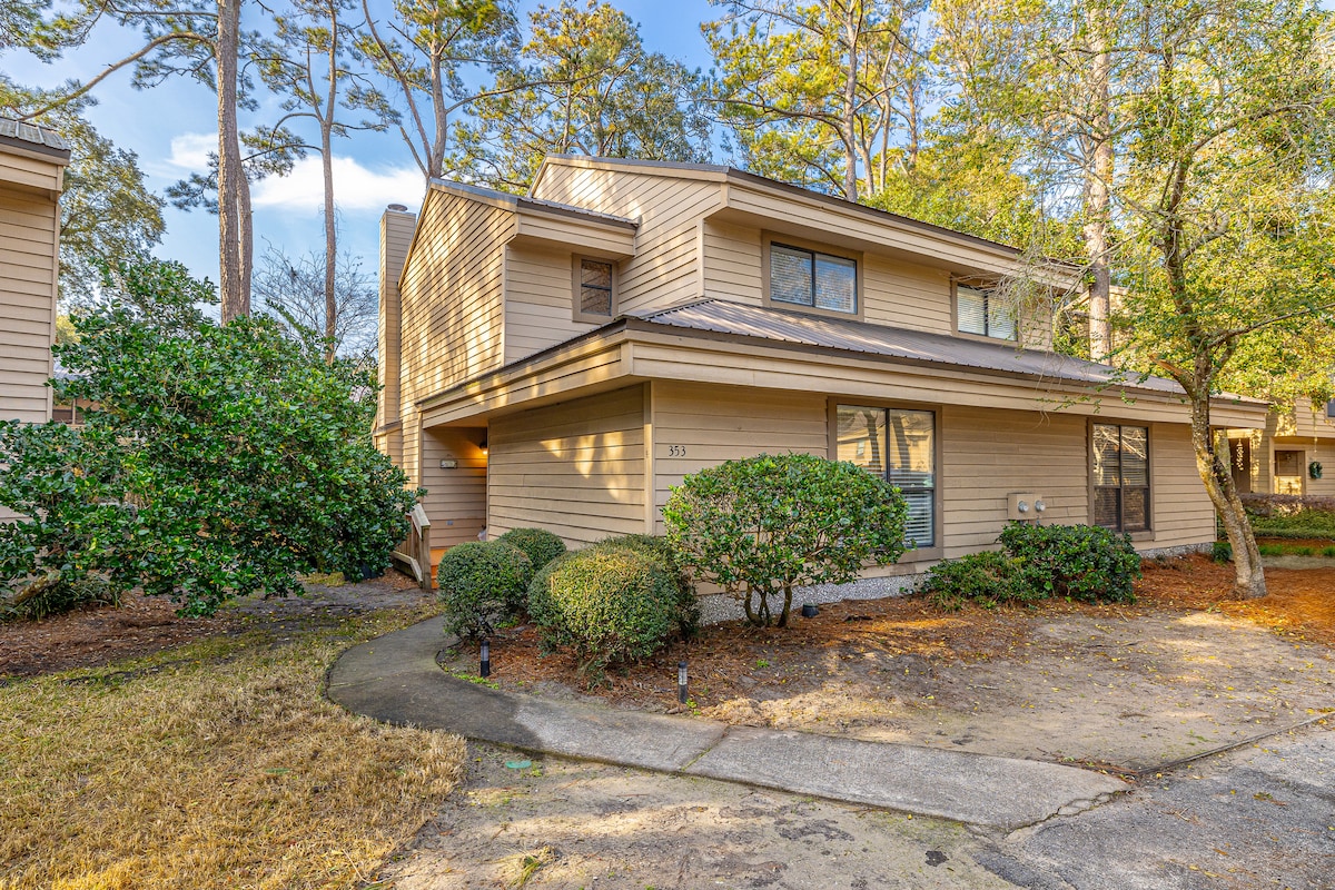 A two-story condo is set in a serene neighborhood, surrounded by trees and greenery. The exterior features light beige siding and a sloped roof, with a pathway leading to the entrance. A well-maintained lawn and shrubs accentuate the inviting entryway.
