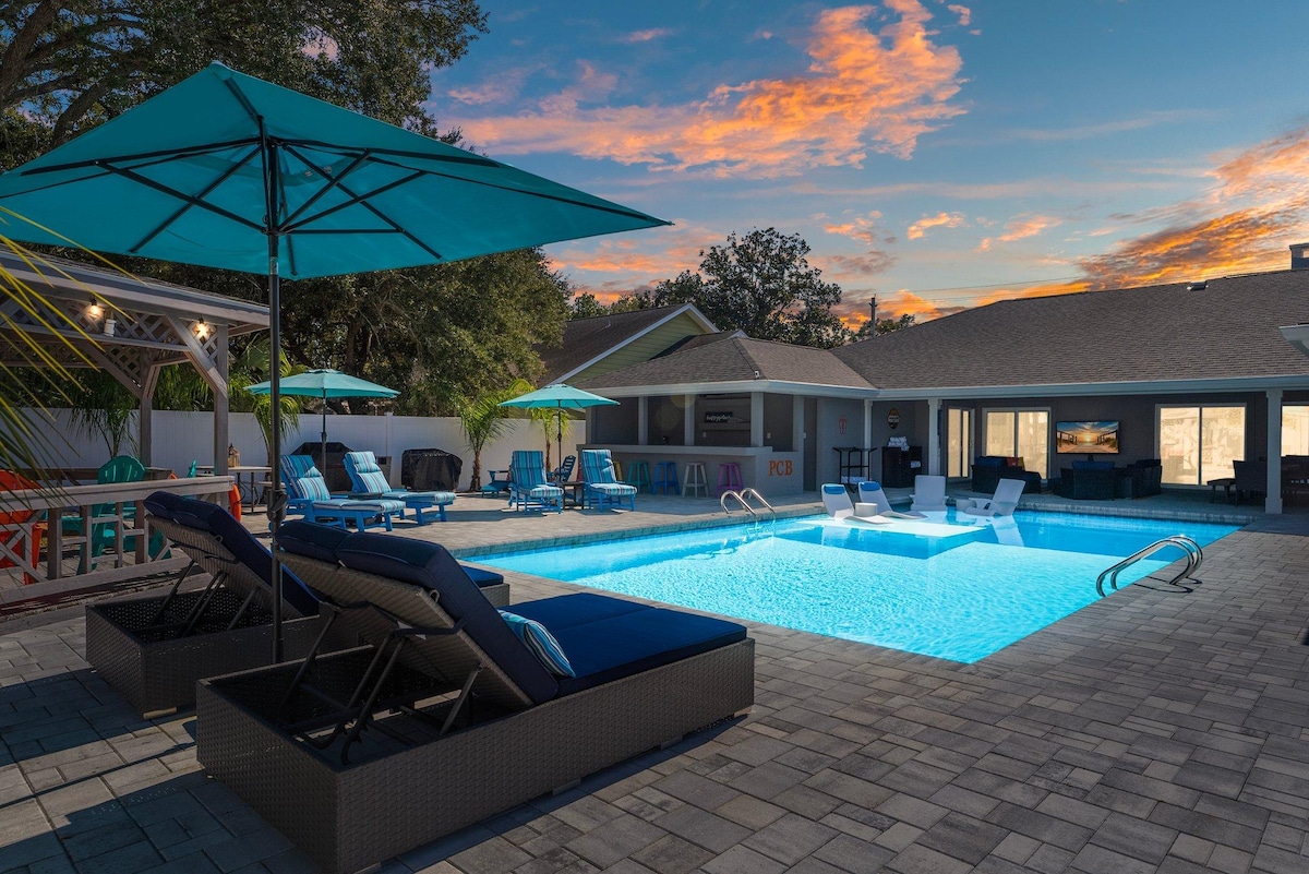 A serene outdoor space features a sparkling private pool illuminated in blue. Lounge chairs and umbrellas provide comfort and shade. The surrounding patio is enhanced by paver stones, and the backdrop includes a glimpse of the home, with large windows inviting natural light.