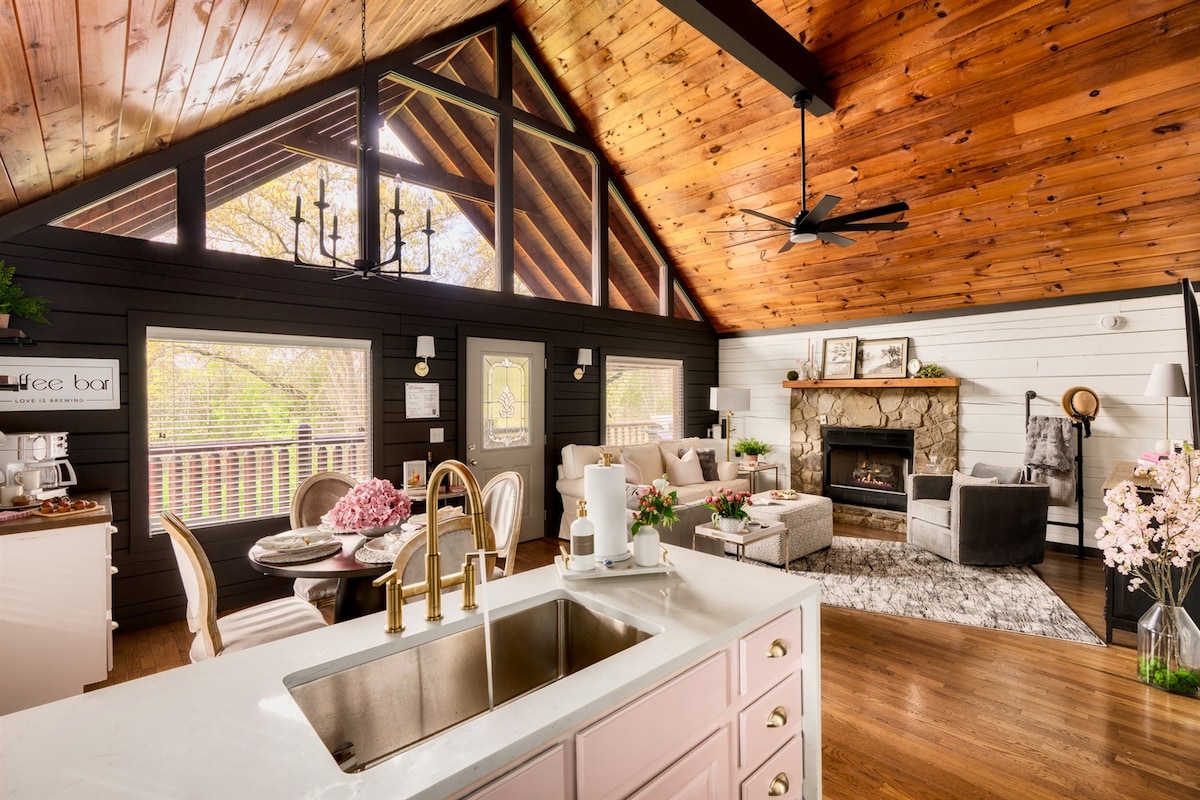 The open-concept living area features high wooden ceilings and large windows allowing ample natural light. A cozy seating arrangement surrounds a stone fireplace, complemented by a well-appointed kitchen with a modern sink. Decorative elements, such as flowers and art, enhance the inviting atmosphere.