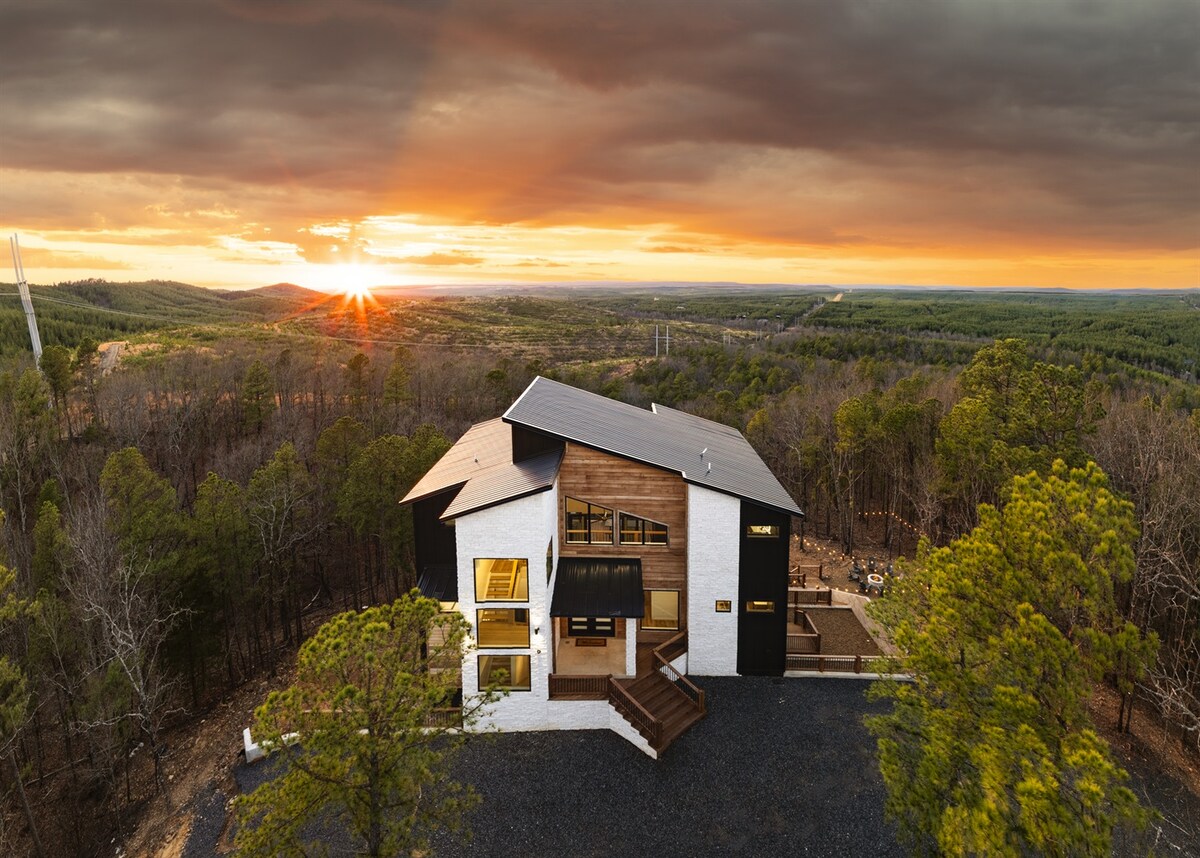 An aerial view of a modern cabin is captured as the sun sets behind distant hills. The cabin’s sleek design features contrasting colors, with expansive windows reflecting the warm light. Surrounded by lush trees, the scene conveys a sense of serenity in nature.