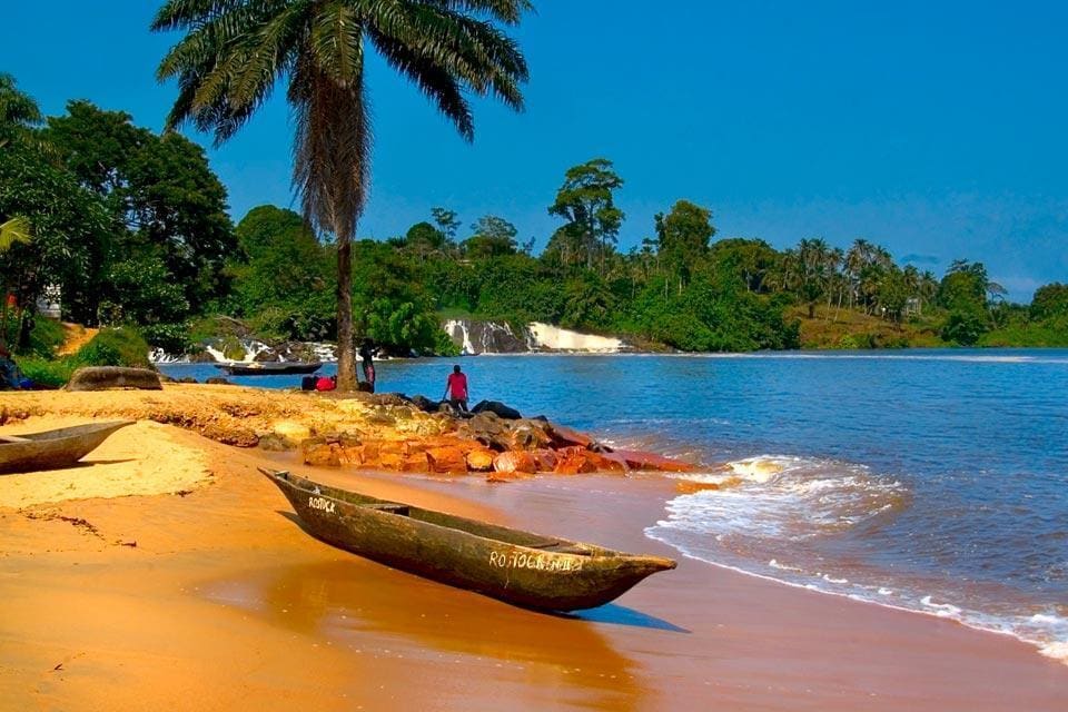 A sandy beach is bordered by lush greenery and gentle waves, with a traditional boat resting on the shore. In the background, soft waterfalls can be seen flowing down, surrounded by trees. Two individuals sit on the rocks, enjoying the serene view.