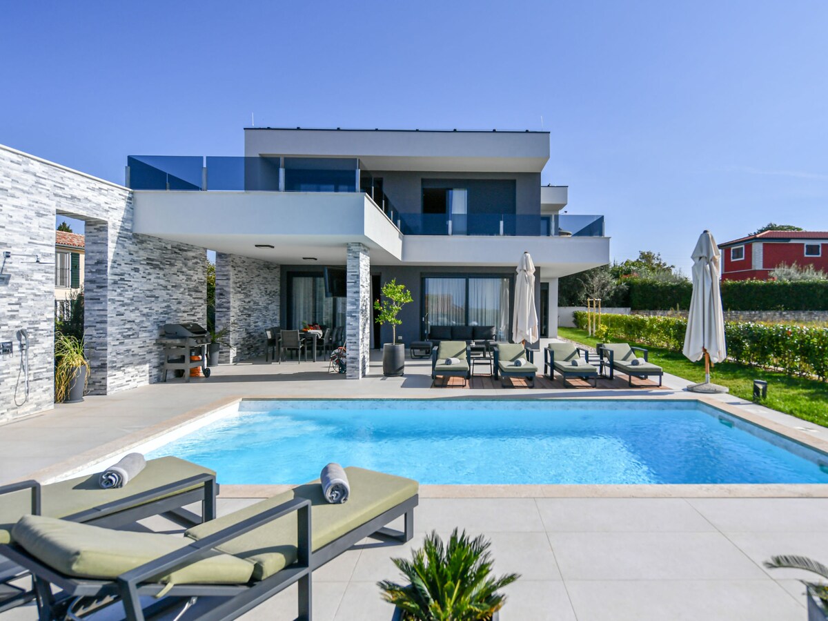 The outdoor area features a heated swimming pool surrounded by lounge chairs. The two-story villa showcases large glass doors opening onto a terrace, with patio furniture and umbrellas under a clear blue sky. Lush greenery and a flower-filled garden are visible in the background.