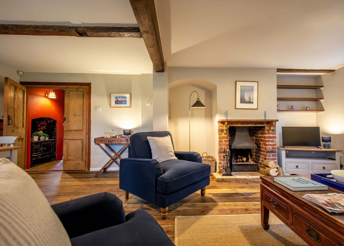 The living room features a cozy seating arrangement with dark blue chairs positioned around a coffee table. A brick fireplace is present, providing a warm focal point. Wood beams across the ceiling and wooden flooring enhance the rustic charm of the space.
