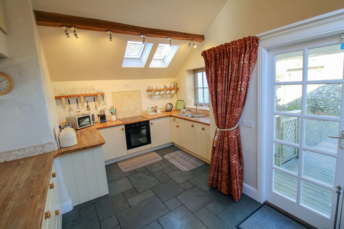 The well-equipped kitchen features wooden countertops and modern appliances, including an oven and kettle. Natural light enters through two skylights, illuminating the slate tiled floor. A door leads to the outdoor patio area, while warm-toned curtains add a touch of coziness to the space.