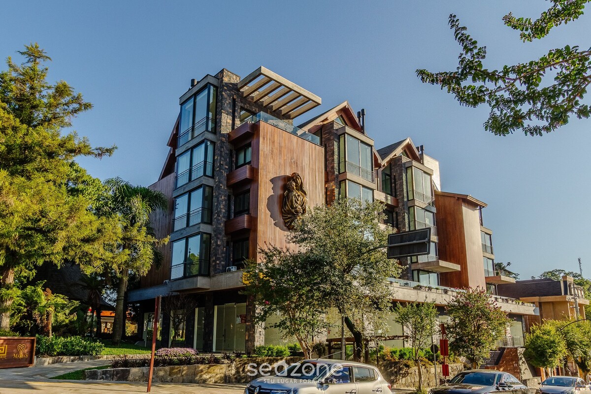 A modern, multi-level building features a combination of stone and wooden elements. Large glass windows reflect the surrounding greenery, while a carved lion's head adorns the facade. The landscaped area includes trees and shrubs, enhancing the building's inviting presence.