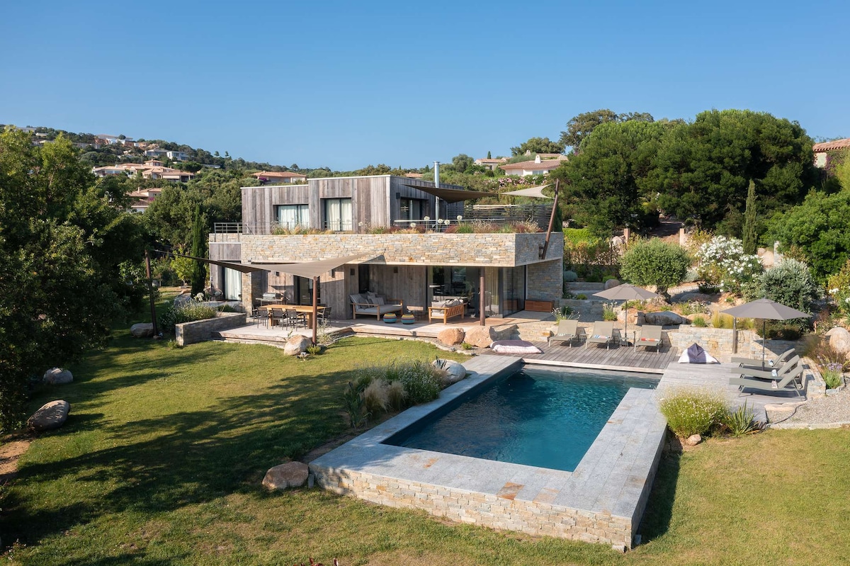 A modern villa is nestled within a landscaped garden, featuring a stone facade and multiple levels. A swimming pool is positioned in the foreground, surrounded by sun loungers and shaded umbrellas, while lush greenery frames the property against a clear blue sky.